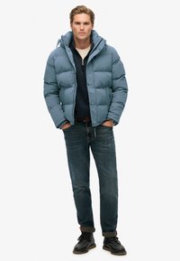 Lys blå vinterjakke med lynlås foran, fast hætte og quiltet tekstur, parret med mørke jeans og sorte støvler.