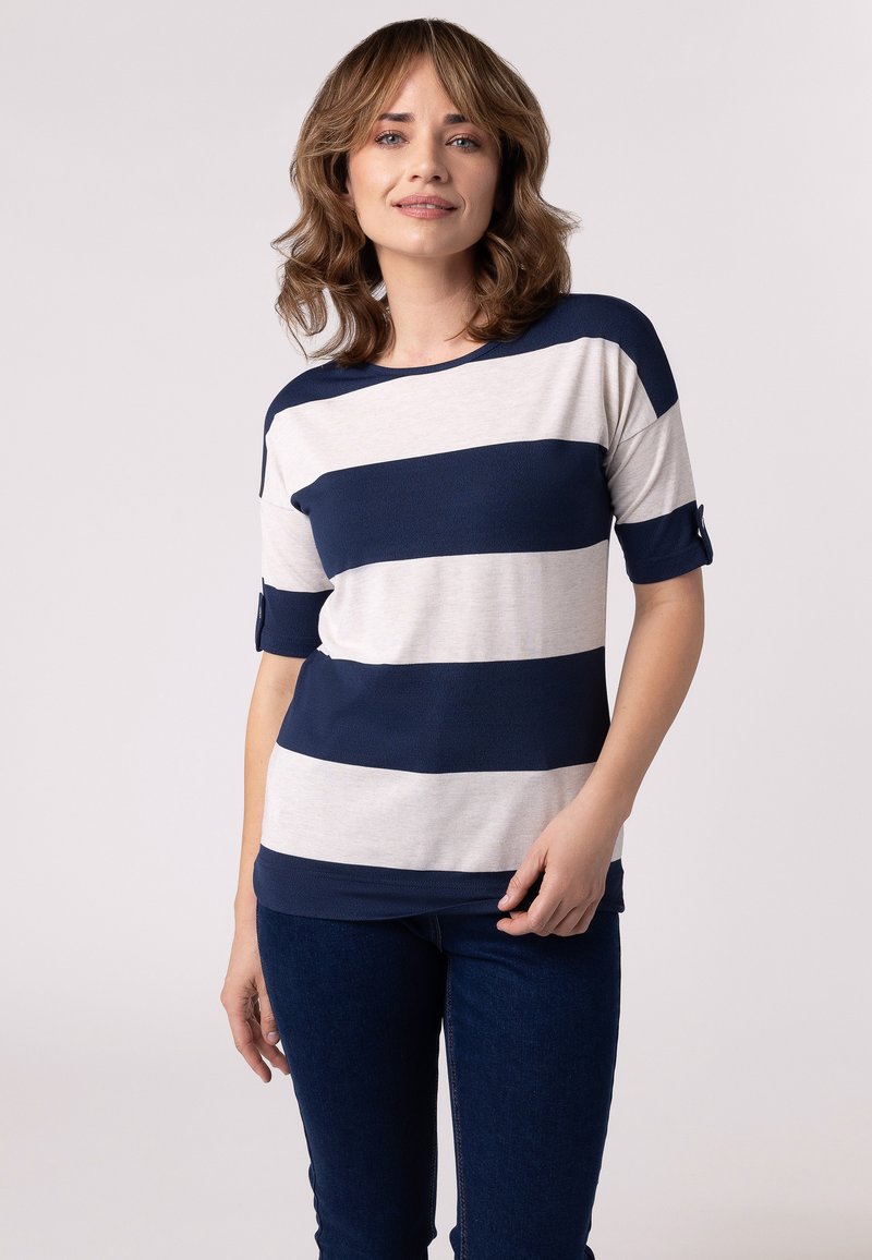 Haut rayé bleu marine et crème à manches courtes, en tissu doux, avec un col rond et des accents de boutons sur les manches. Porté avec un jean bleu foncé.