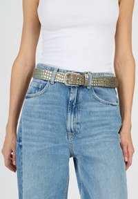 Ruskea kudottu vyö metallisine yksityiskohtineen, jossa on teksturoitu ruudukkomalli ja hopeinen suorakulmainen solki, käytettävä vaaleansinisten farkkujen päällä.