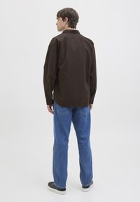 Joven de pie de espaldas, vistiendo una camisa de manga larga marrón oscuro, jeans azules y zapatillas negras con suelas blancas.