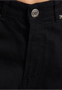 Jean en denim noir avec une coupe ajustée ; présente un bouton métallique gravé "DEF", des détails de coutures verticales et une texture lisse.
