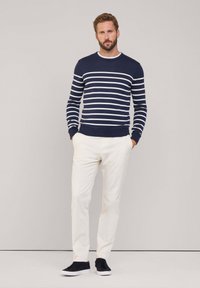 Maglione a righe blu navy con strisce bianche, colletto a girocollo, abbinato a pantaloni bianchi e sneaker scure. Texture liscia, design casual.