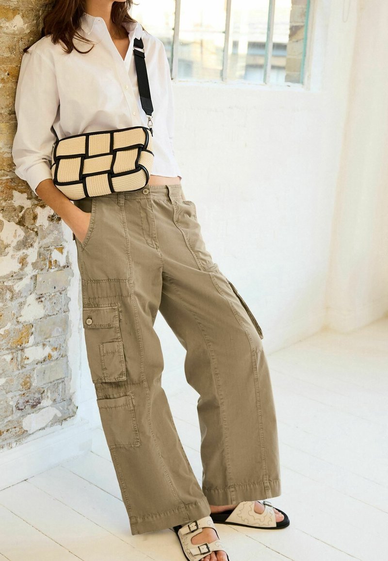 Personne portant un pantalon cargo beige, une chemise blanche boutonnée, des sandales blanches et portant un sac bandoulière tissé beige et noir.