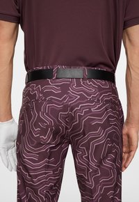 Spodnie sportowe w ciemnym fioletowym kolorze z geometrycznym wzorem w jaśniejszym fioletowym. Posiadają czarny pasek, boczne kieszenie i teksturowany materiał.