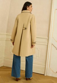 Femeie purtând o geacă bej de tip trench cu pliuri la spate și bretele cu nasturi, blugi albastre cu picioare largi și pantofi maro cu toc, în interior.