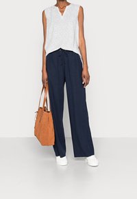 Top bianco leggero senza maniche con motivo grigio, abbinato a pantaloni larghi blu navy, sneakers bianche e una borsa in suede marrone.