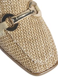 Chaussure slip-on beige tissée avec un bout pointu, dotée d'un accent en métal doré et d'une semelle lisse couleur tan. Le tissu texturé offre un aspect naturel.