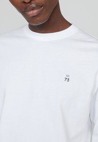 Baltos ilgomis rankovėmis marškiniai, pagaminti iš medvilnės, su apvalia iškirpte. Išskirtinė pilka logo su užrašu "M 73", atspausdinta ant krūtinės. Lygi tekstūra.