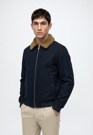 Homme portant une veste bleu marine avec un col en polaire marron et un pantalon beige, debout les mains dans les poches devant un fond uni.