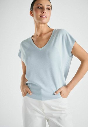 Woman wearing a light blue short-sleeve V-neck sweater and white pants with hands in pockets, looking to her left against a plain background.