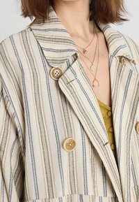 Pruhovaná bunda v krémové, žluté a námořnické barvě; má velké knoflíky a vykrojený límec; vyrobena z texturované látky. Přes žluté tričko.