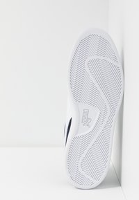 Zapatilla blanca con suela de goma texturizada que presenta un patrón de espiga, logo de la marca y detalles en azul oscuro en los paneles laterales.