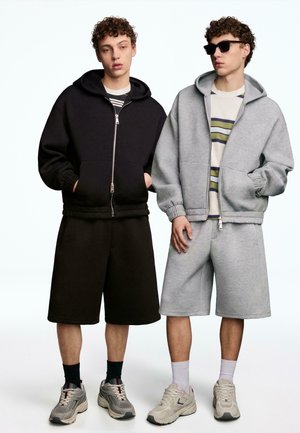 Twee mannen dragen capuchonvesten met rits en lange shorts; één in het zwart met een gestreept shirt, één in het grijs met een gestreept shirt en een zonnebril.