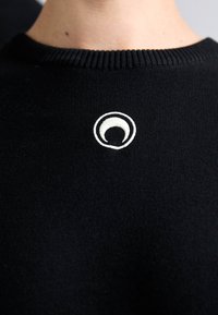 Fekete pulóver bordázott nyakrésszel. Fehér hímzett, kerek logó található a mellrészen. Simaság és minimalista dizájn jellemzi.