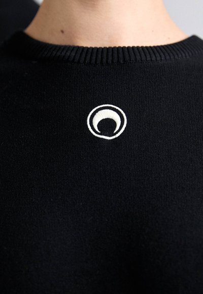 Črna majica s pletenim ovratnikom. Ima bel vezen krožni logotip na prsih. Gladka tekstura, minimalističen dizajn.