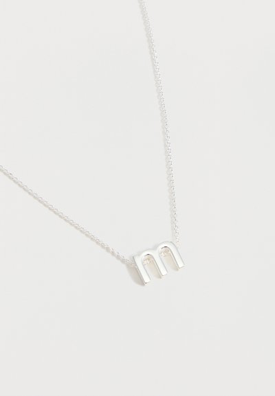 Collier en argent avec un pendentif en minuscule « m » sur une chaîne délicate, présenté sur un fond blanc uni.