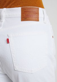Pantalones vaqueros de mezclilla blanca con una etiqueta de cuero marrón de Levi Strauss, detalles de costura visibles y un acento rojo en el bolsillo trasero.