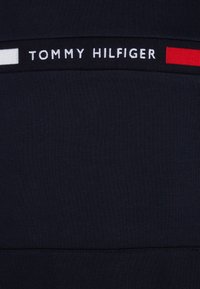 Nærbillede af marineblåt stof med et sort bånd med hvid tekst "Tommy Hilfiger" og hvide og røde rektangulære farveblokke på hver side.