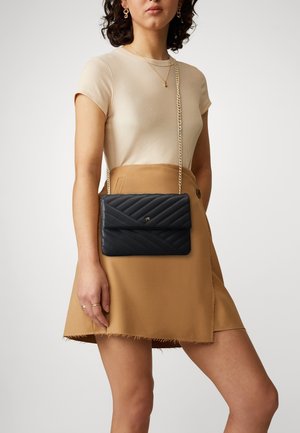 Kvinde i beige ribstrikket skjorte og tan nederdel bærer en sort quiltet crossbody-taske med guldkæde og lille guldemblem.