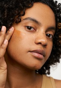 Fond de teint liquide marron appliqué sur la joue, à côté d'une peau lisse et sans imperfections. Des cheveux bouclés encadrent le visage, doucement éclairé.