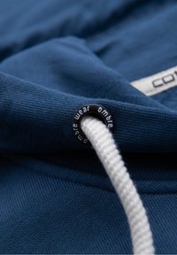 Gros plan d'un cordon blanc passant à travers un œillet en métal noir sur un tissu bleu avec une étiquette de marque partiellement visible.