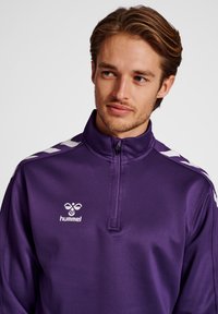 Fioletowa kurtka sportowa z białymi akcentami, zamek błyskawiczny do jednej czwartej, z logo na piersi. Gładka faktura i lekk materiał.