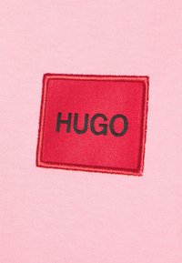 Różowy materiał z czerwoną prostokątną łatą z czarnym haftowanym napisem "HUGO". Tkanina wygląda na gładką z lekkim rozciągnięciem.