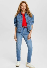 Jeansjacke mit lockerer Passform, blauer Farbe, mit Fronttaschen; kombiniert mit einem roten Hemd, hoch taillierten blauen Jeans und weißen Sneakers.