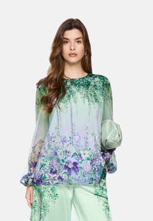 Frau trägt eine floral grün-lila Langarmbluse mit passenden Hosen und hält eine hellgrüne, strukturierte Clutch.
