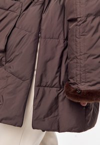 Chaqueta acolchada marrón con textura acolchonada, que presenta puños de piel y bolsillos con cremallera. El tejido parece suave y ligero.