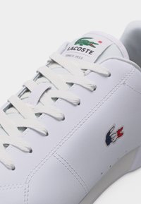 Baskets blanches Lacoste avec lacets blancs, logo crocodile vert sur la languette et emblème crocodile rouge, blanc et bleu sur le côté.