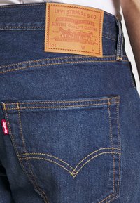 Tummansiniset denimfarkut, joissa on vaaleanruskea nahkainen merkkilaatta, oranssit tikit ja punainen Levi's-tunniste takataskussa.