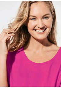 Felle roze T-shirt met een ronde halslijn, gemaakt van gladde stof. Onderwerp heeft lang, golvend haar en toont een lichte, natuurlijke glimlach.