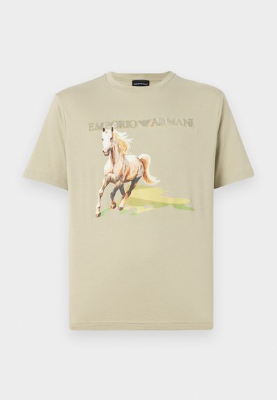 T-shirt en coton beige avec un graphique de cheval imprimé. Le logo "Emporio Armani" est au-dessus du design dans une couleur claire. Manches courtes, col rond.