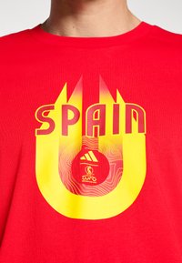 Červené bavlněné tričko s výraznou žluto-červenou grafikou s nápisem "ŠPANĚLSKO" a kruhovým logem pod ním. Kulatý výstřih, krátké rukávy.