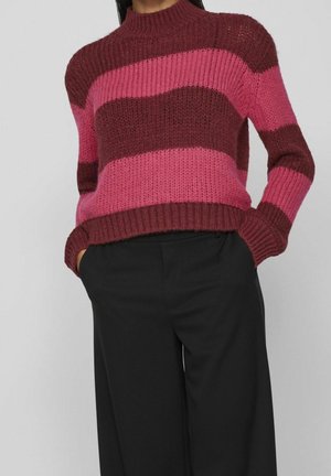 Person iført en ribstrikket sweater med brede horisontale vinrøde og lyserøde striber kombineret med løse sorte bukser, hænderne i lommerne.