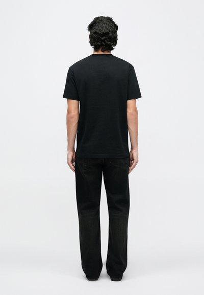 Vīrietis stāv ar muguru pret skatītāju, valkā vienkāršu melnu īsroku kreklu un melnas džinsu bikses uz balta fona.