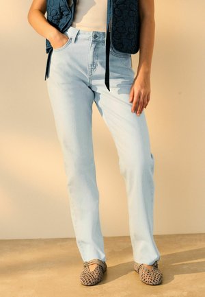 Personne portant un jean taille haute bleu clair, un haut blanc, un gilet bleu foncé et des chaussures tissées beiges, debout sur un sol clair.