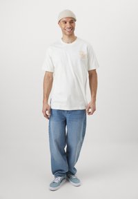 Vit bomullströja med orange tryckt text "1956" på bröstet. Kombinerad med ljusblå baggyjeans och teal sneakers.