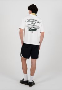 Hvit t-skjorte med korte ermer med et grafisk trykk av en bil og tekst på baksiden, kombinert med svarte shorts og svarte sko.