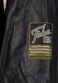 Sort læderjakke med et grønt og hvidt stofmærke, hvor "Fubu" står med store kursiv bogstaver og "05" nedenunder, plus mindre tekstdetaljer.