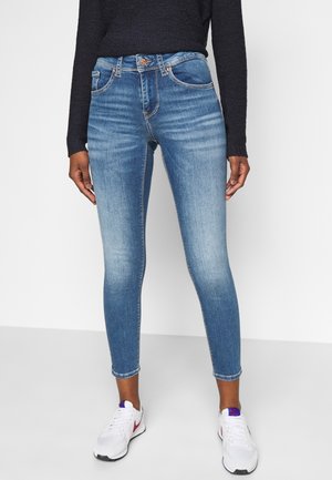 Vrouw die vervaagde blauwe skinny jeans en witte sneakers draagt, staand tegen een effen lichte achtergrond, met een zwart shirt met lange mouwen.