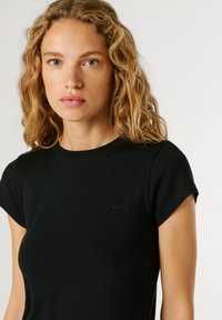 Junge Frau mit lockigem blondem Haar, die ein schlichtes schwarzes T-Shirt mit kurzen Ärmeln trägt und vor einem weißen Hintergrund nach vorne schaut.