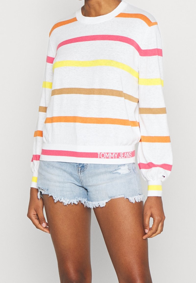 Femme portant un pull blanc à manches longues avec des rayures horizontales orange, rose, jaune et marron, associé à un short en jean bleu clair déchiré.