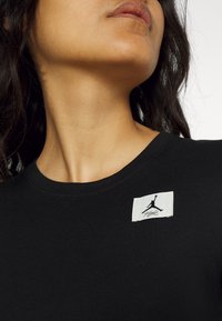 T-shirt nera con collo rotondo, caratterizzata da un'etichetta in tessuto bianco che mostra un logo con una figura che salta e del testo, posizionata sul petto.