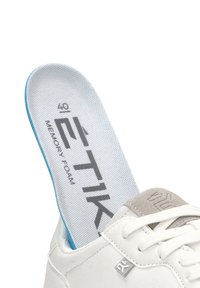 Zapatilla deportiva blanca con un acento de ante gris y cordones. Plantilla de espuma viscoelástica azul visible, con superficie texturizada y marca impresa.