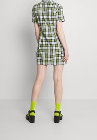Mujer vestida con un vestido corto de cuadros verdes y blancos, calcetines verdes neón y sandalias de tacón negro, de pie contra un fondo liso claro.