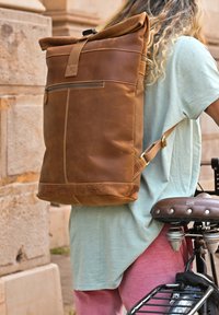 Hnedý kožený ruksak s rolovateľným vrchom, prednou kapsou, nastaviteľnými popruhmi a kontrastným prešívaním. Nosený osobou vedľa bicykla.