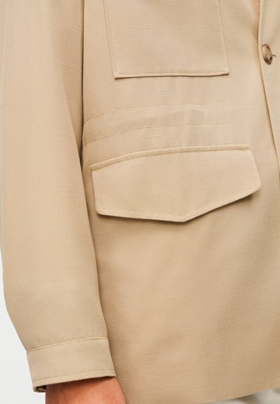 Μπεζ μακρυμάνικο πουκάμισο με λεία υφή, δύο τσέπες στο στήθος, κούμπωμα με κουμπιά και διπλωμένα μανίκια για μια δομημένη εμφάνιση.