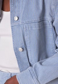 Giacca a righe blu chiaro con bottoni bianchi, dotata di una tasca sul petto e vestibilità leggermente ampia. La texture del tessuto è liscia e tessuta.